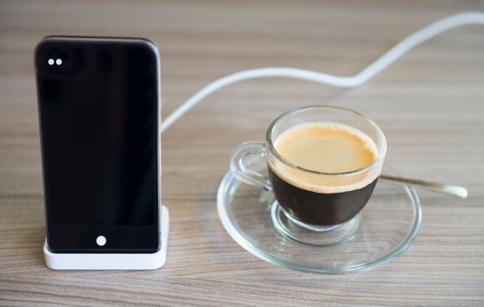 Celular carregando em suporte de mesa para celular branco ao lado de uma xícara de café.