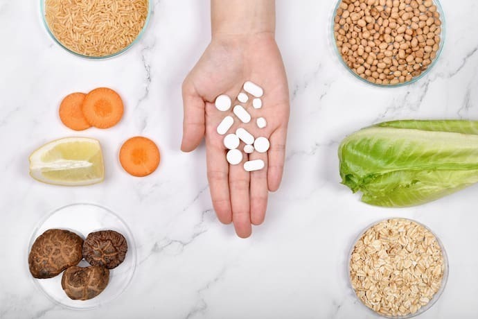 Mão segurando polivitamínico com vários legumes e verduras ao redor.