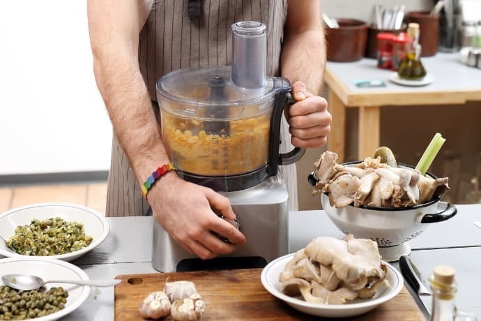 Homem de avental preparando um prato de vegetais com processador.