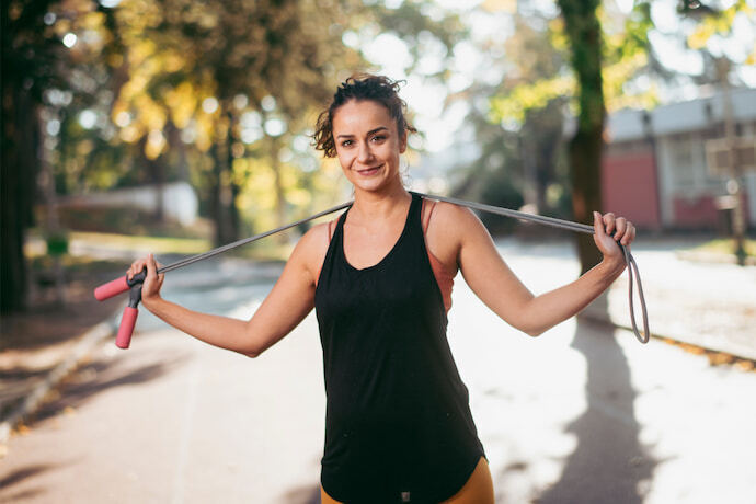 Mulher sorridente com regata preta, segurando uma corda de pular ao ar livre.