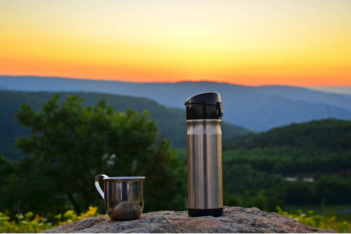 Garrafa térmica e caneca em um fundo de paisagem natural.